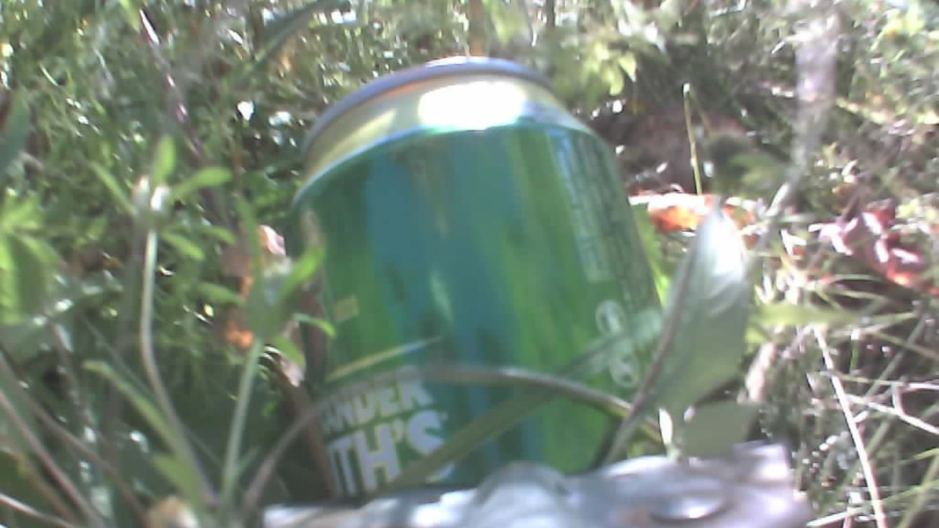 A green beer can gripped in the litter picker’s jaws among weeds, seen from the camera mounted above the jaws.