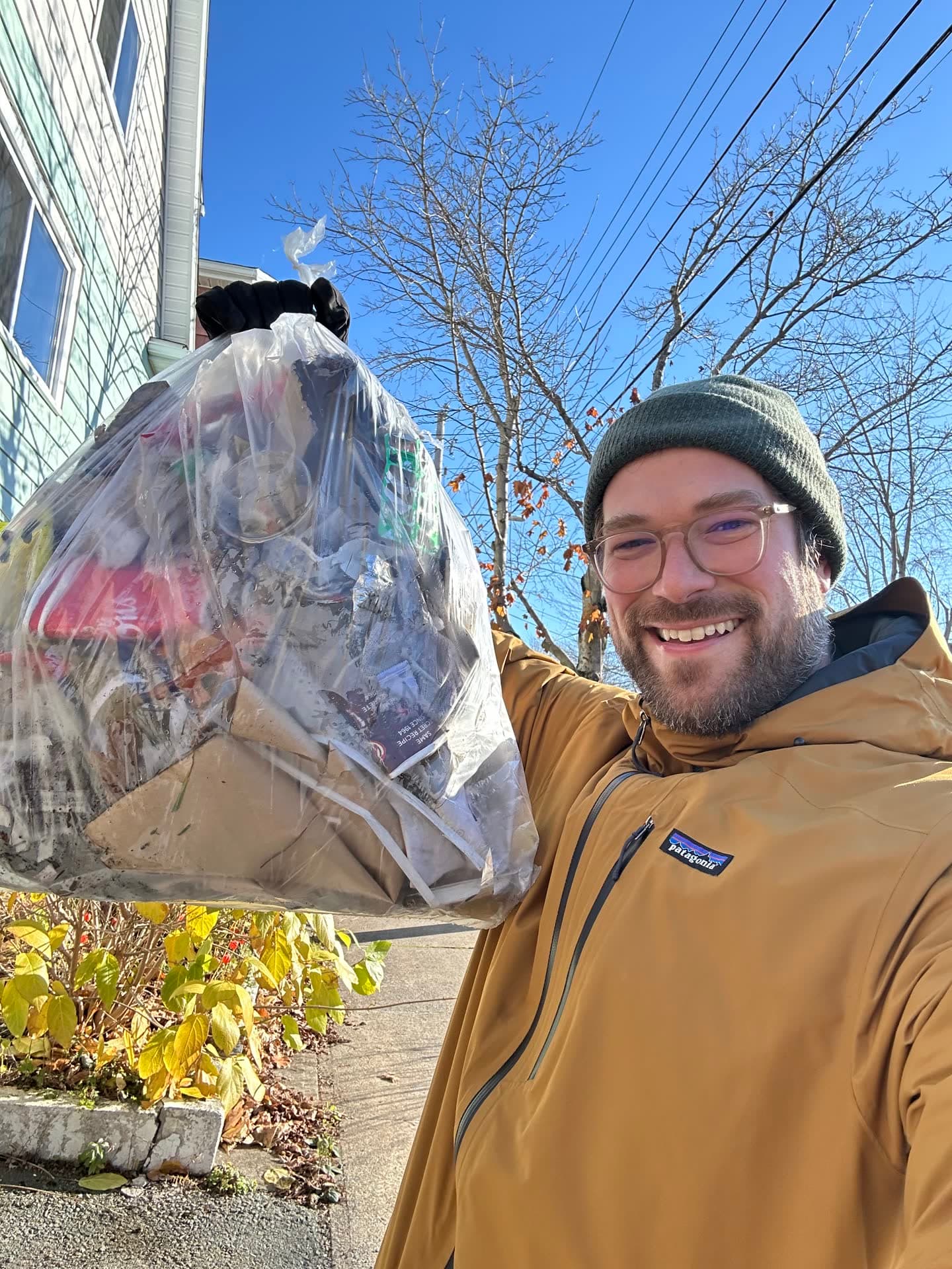 Jesse in a toque on a sunny Halifax sidewalk, grinning and holding up a full clear bag of litter.