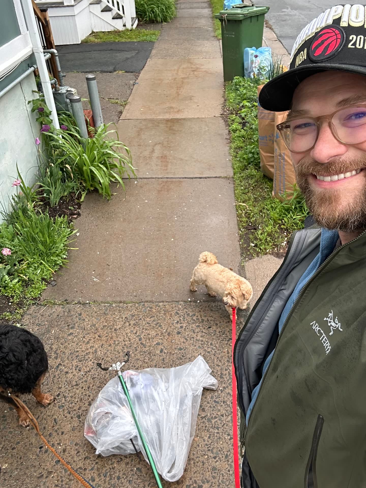 Jesse on a sidewalk with two small dogs on leads, a litter picker and a bag of trash at his feet.