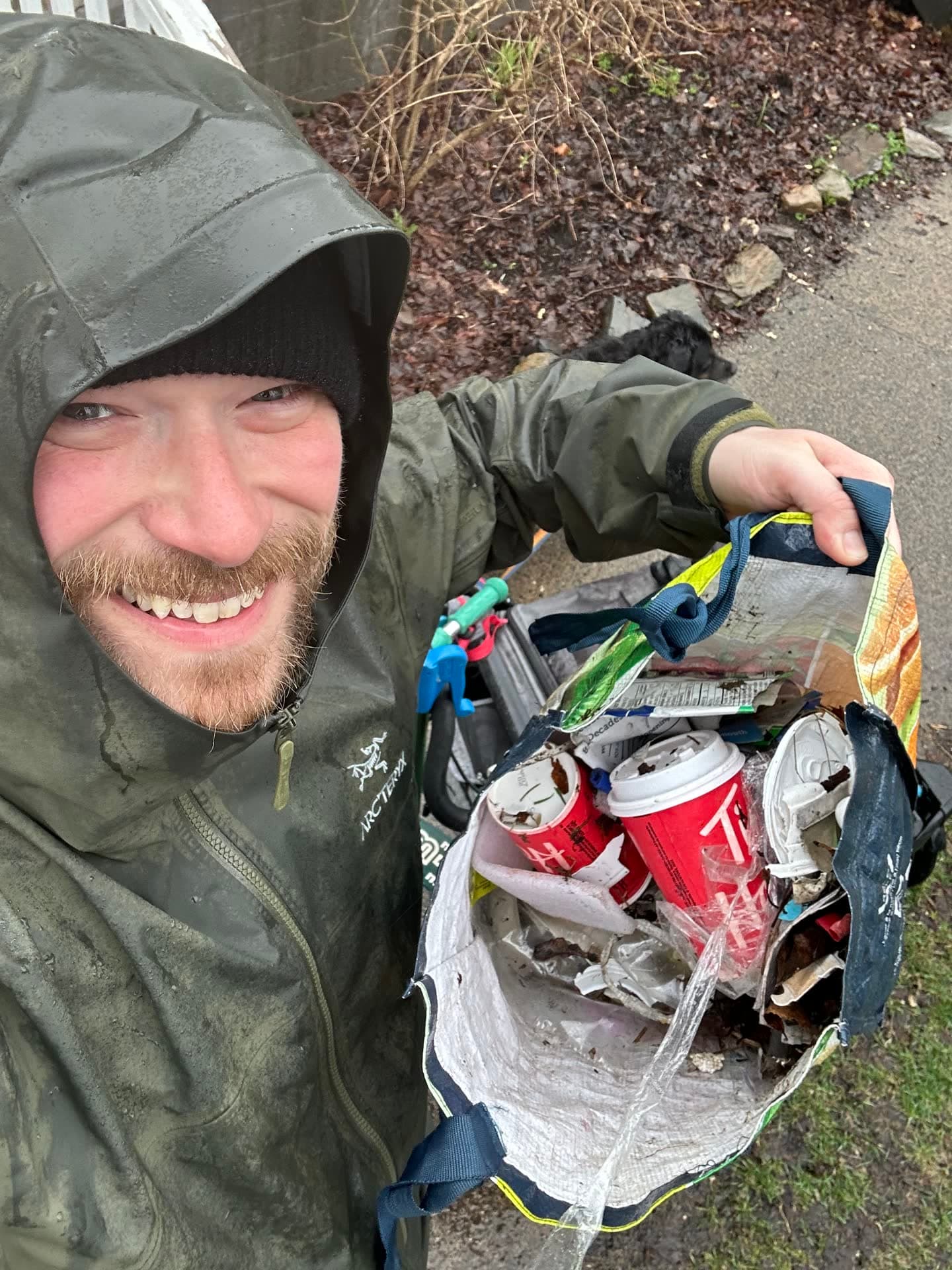 Jesse in a rain jacket with the hood up, smiling in the rain and holding open a bag full of cans and wrappers.